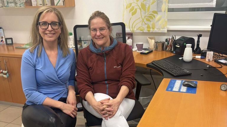 Two women in glasses sit in an office, one wearing a blue shirt and the other a maroon hoodie. The woman on the right is smiling, and the woman on the left is looking at her. Behind them is a desk with a keyboard, cup, and scissors.