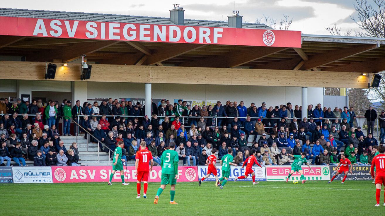 A soccer game in progress at the Siegendorf stadium with players wearing green and red jerseys. The audience is seated on the bleachers.