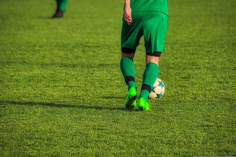 Ein Fußballspieler in Grün kickt einen Ball auf einem Feld mit einem Schatten auf dem Gras.