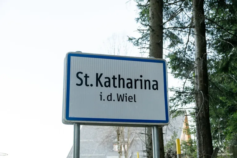A white sign with blue borders displays 'St. Katharina i.d. Wiel' against a background of trees and a gray sky.