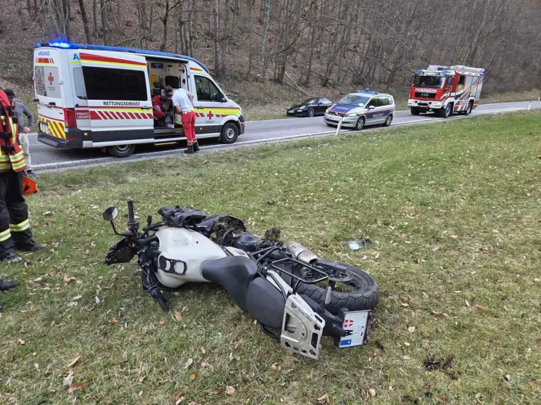 An overturned motorcycle is beside the road, with an ambulance, a police car, and a fire truck parked nearby. A person in red pants stands by the open ambulance door.