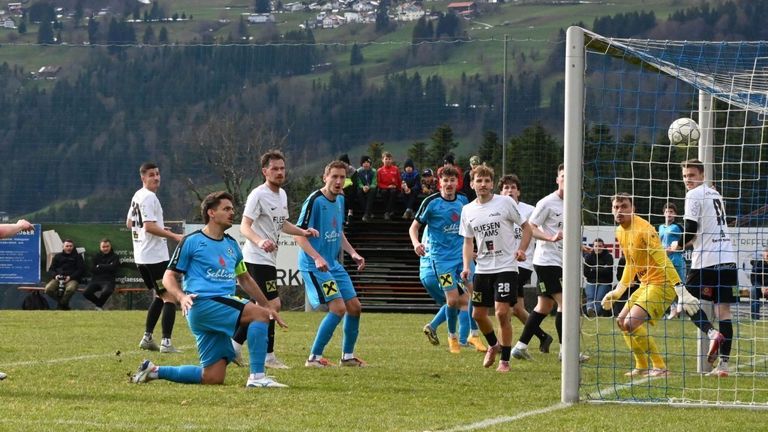 Football players wearing blue uniforms are running on a grassy field. One of them is kneeling down. Behind them, a net is fixed on a pole. Spectators are watching from the stands.
