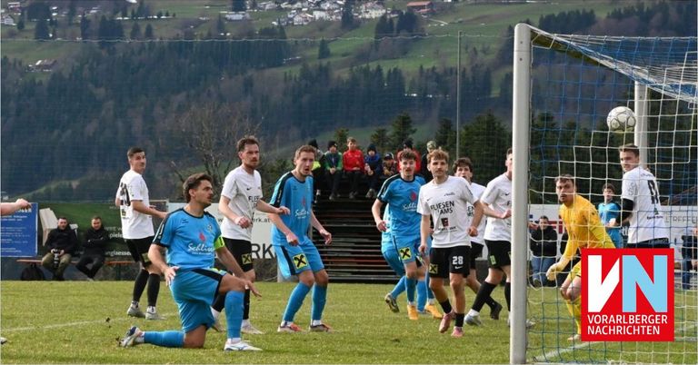 Soccer players in blue uniforms run towards the goal. One player has fallen to the ground. Spectators watch from the stands.