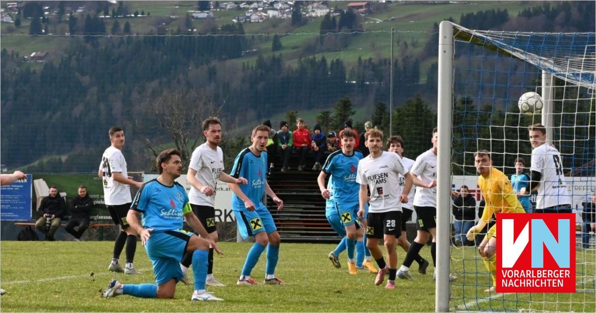 Soccer players in blue uniforms run towards the goal. One player has fallen to the ground. Spectators watch from the stands.