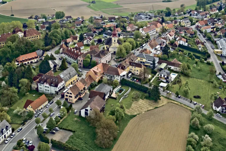 Ein Luftbild einer europäischen Ortschaft mit Häusern und einer Kirche, umgeben von Bäumen und grünen Feldern, mit einer Straße und geparkten Autos.