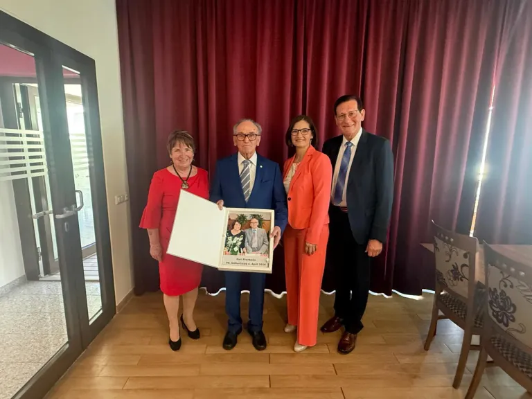 Vier Menschen stehen vor einem roten Vorhang und lächeln. Ein Mann hält ein Fotoalbum. Die Frau links trägt ein rotes Kleid und der Mann rechts einen blauen Anzug.