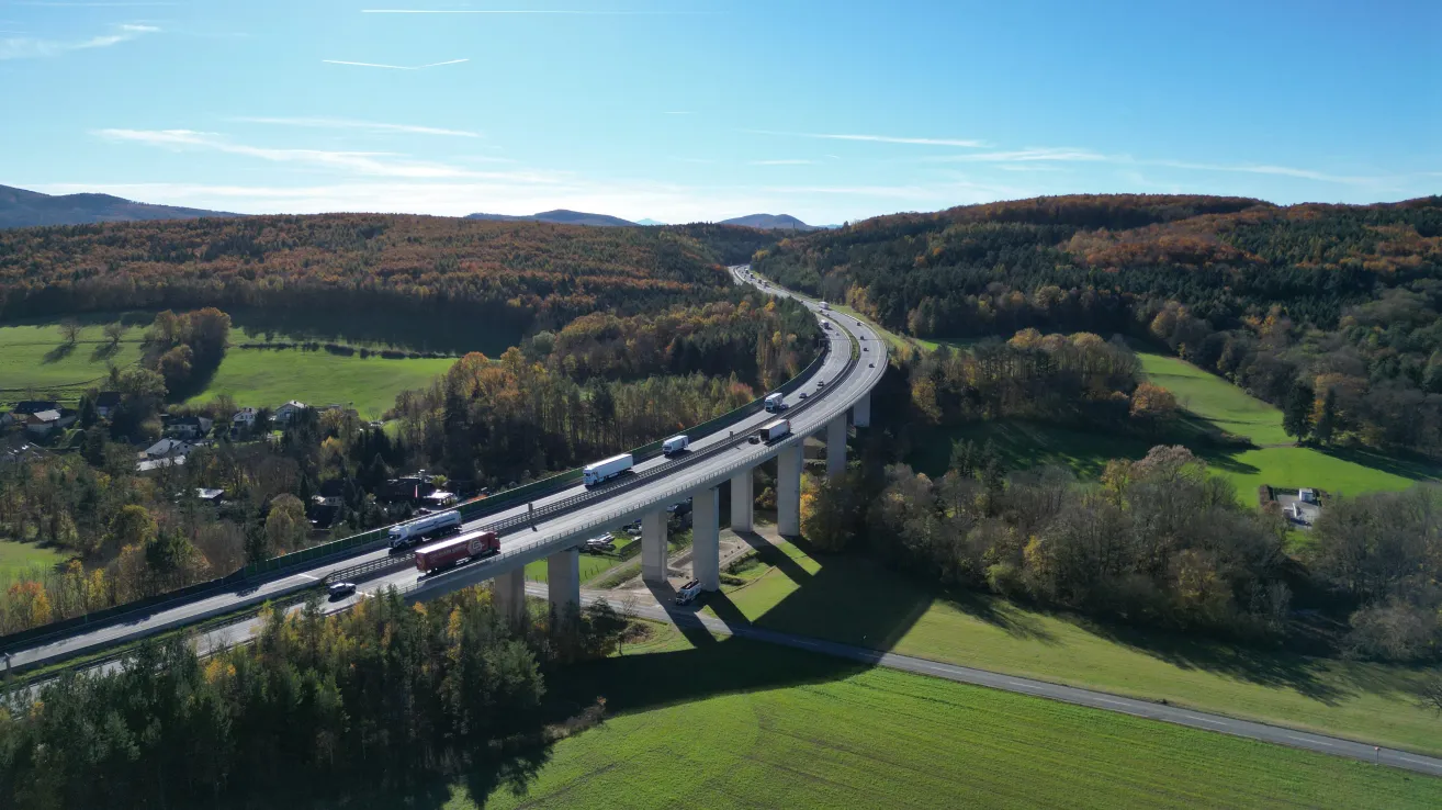 Luftaufnahme einer Autobahn mit mehreren Fahrzeugen, darunter Lastwagen, die auf einer Brücke über einem grünen Tal fahren, unter einem blauen Himmel.