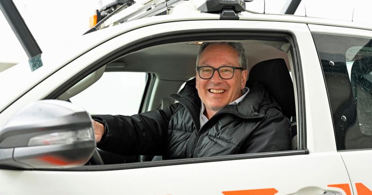 A smiling man wearing glasses sits in the driver's seat of a white vehicle with a black roof rack and an orange rear light.