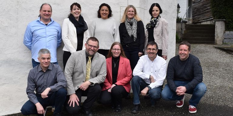 A group of people are posing for a photo. They are standing and sitting in front of a building. They are all smiling and looking in the same direction.