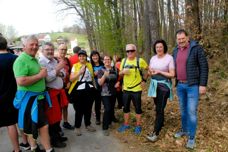 A group of hikers, dressed in casual outdoor gear, stand together on a forest path, smiling and holding drinks. Trees and greenery surround them, with houses visible in the distance.