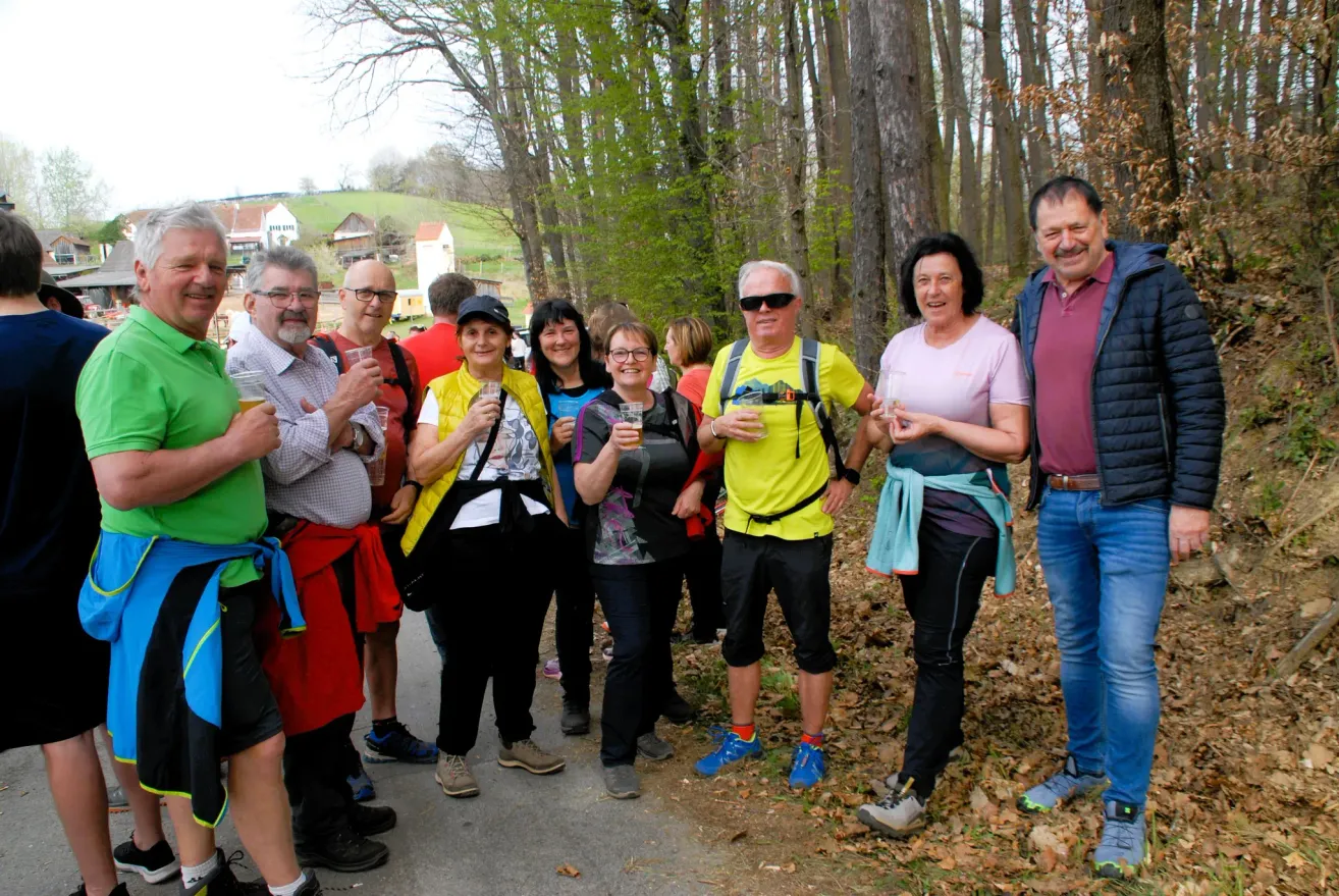 A group of hikers, dressed in casual outdoor gear, stand together on a forest path, smiling and holding drinks. Trees and greenery surround them, with houses visible in the distance.
