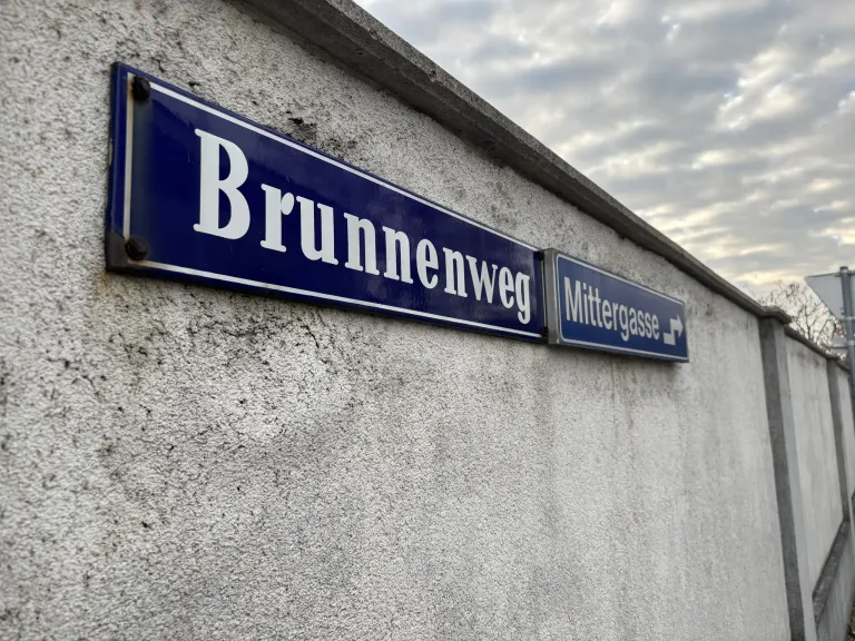Zwei blaue Straßenschilder an einer Wand, eines mit der Aufschrift 'Brunnweg' und das andere 'Mittergasse' mit einem Pfeil nach rechts. Im Hintergrund ist ein bewölkter Himmel zu sehen.