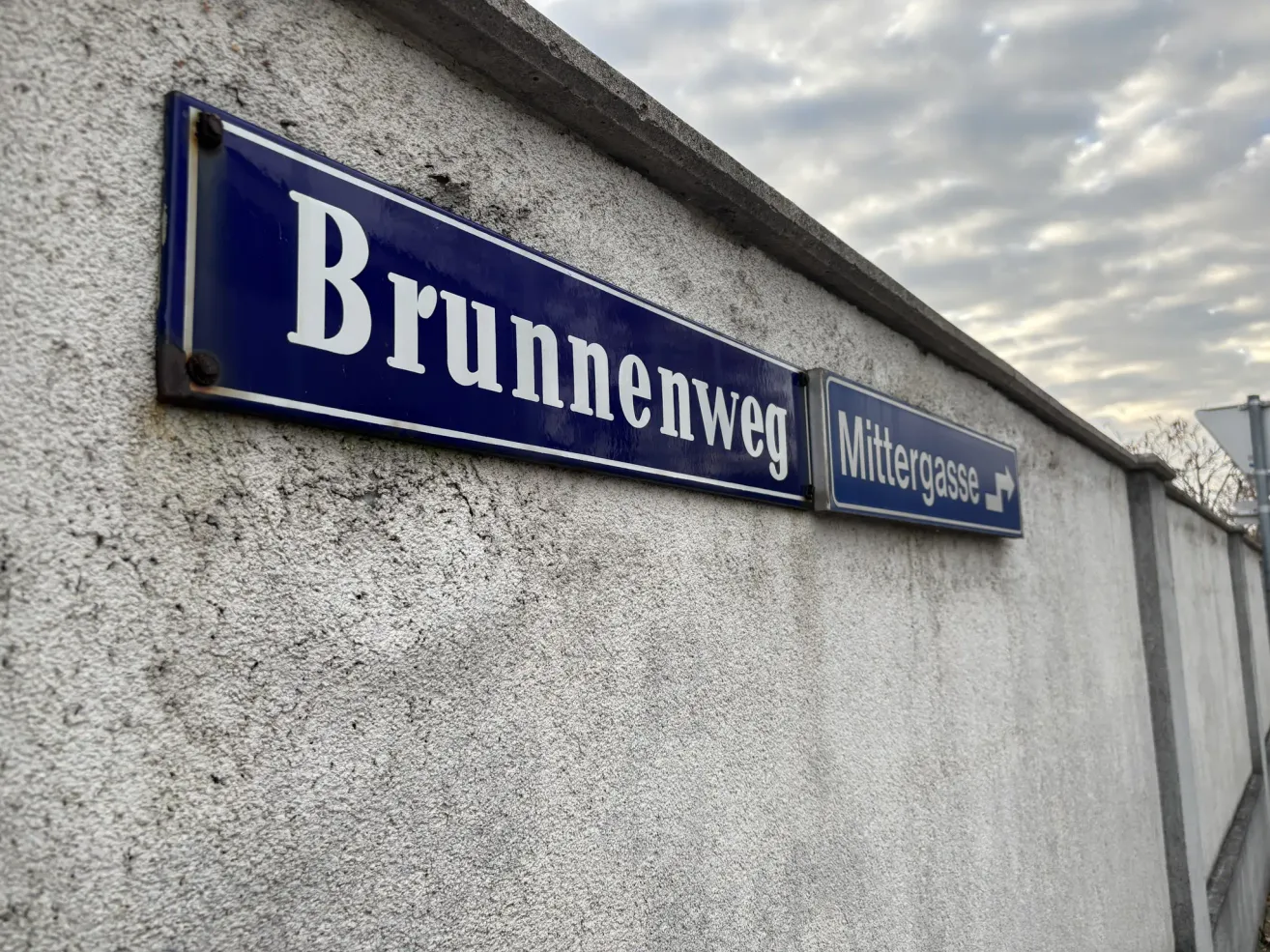 Zwei blaue Straßenschilder an einer Wand, eines mit der Aufschrift 'Brunnweg' und das andere 'Mittergasse' mit einem Pfeil nach rechts. Im Hintergrund ist ein bewölkter Himmel zu sehen.