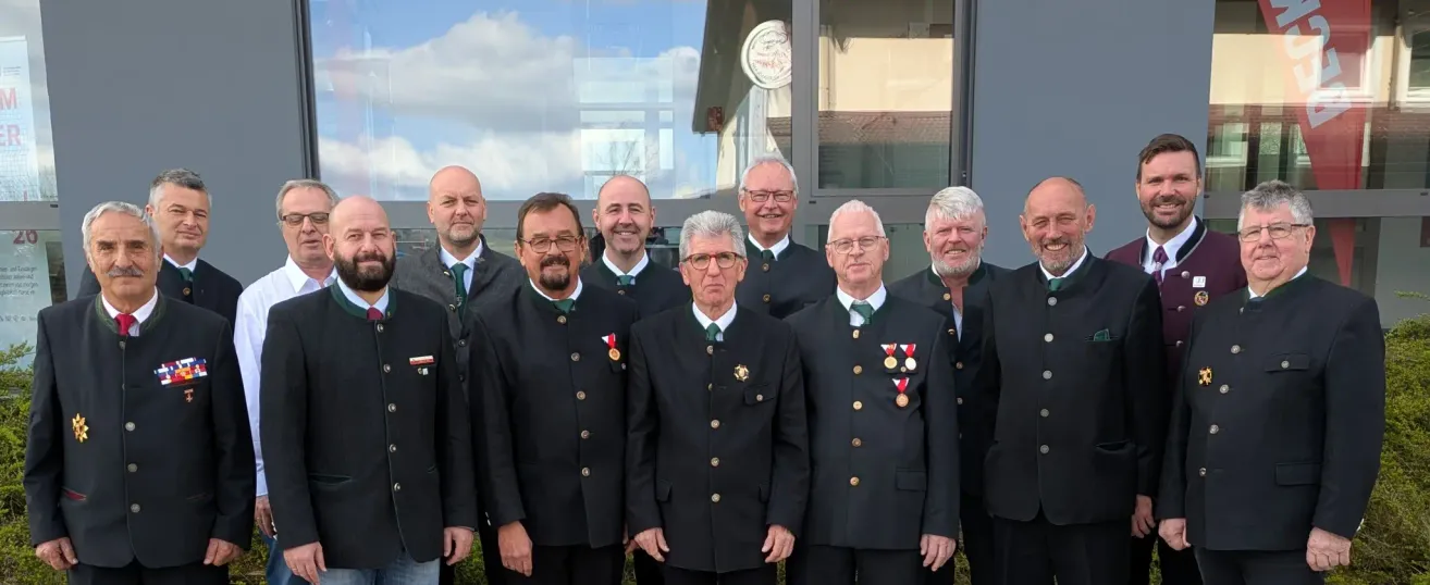 A group of men in formal attire, possibly a club or organization, posing for a photo with a smiling expression. They are standing in front of a building with glass windows.