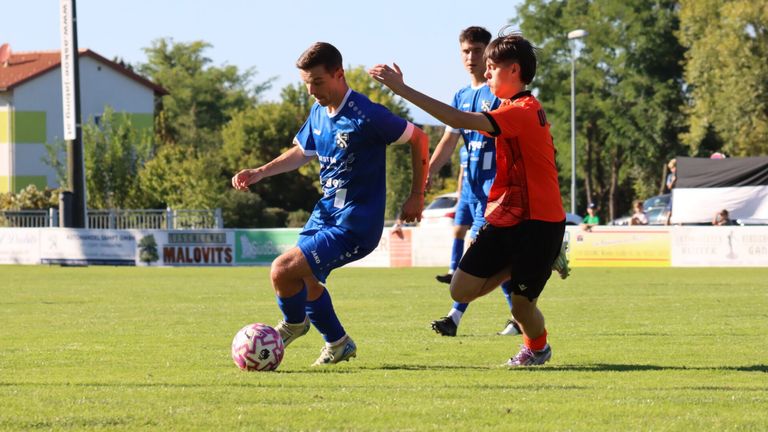Ein Fußballspiel mit Spielern in blauen und orangefarbenen Trikots. Ein Spieler in Blau tritt einen Fußball, während ein anderer in Orange versucht, ihn zu blockieren. Dahinter steht ein Auto in der Nähe von Bäumen.