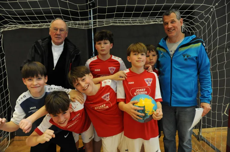 A group of boys in red jerseys with names on them are posing for a photo with a globe. Two men in different colored jackets stand behind them near a net.