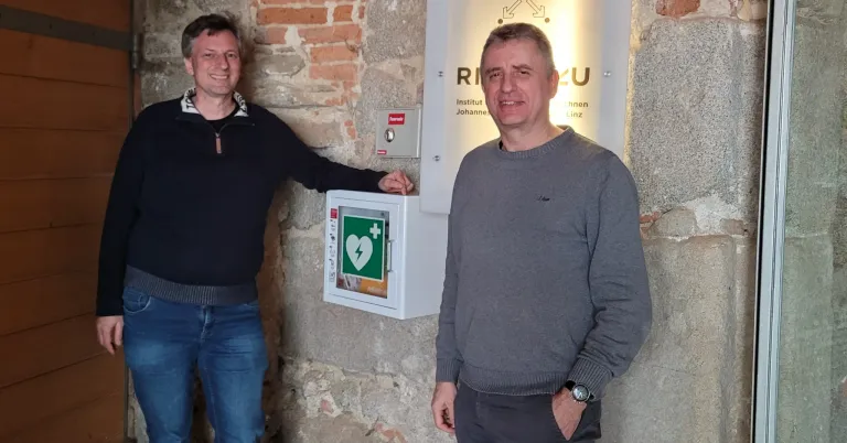 Two men stand beside a defibrillator mounted on a stone wall. Both are smiling.