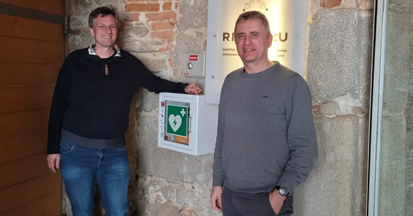 Two men stand beside a defibrillator mounted on a stone wall. Both are smiling.