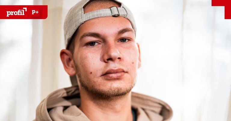 Ein Mann mit Bart und Baseballcap schaut direkt in die Kamera. Er trägt eine beigefarbene Kapuzenjacke und hat einen roten Fleck auf der Stirn.