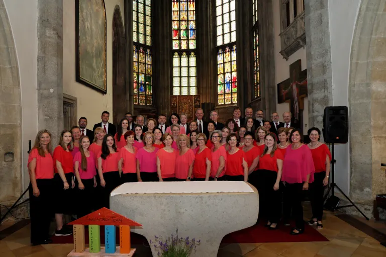 A choir group stands in a church, wearing pink tops and black bottoms. Behind them, men in suits and ties stand in front of stained glass windows. The choir is positioned in front of a white altar.