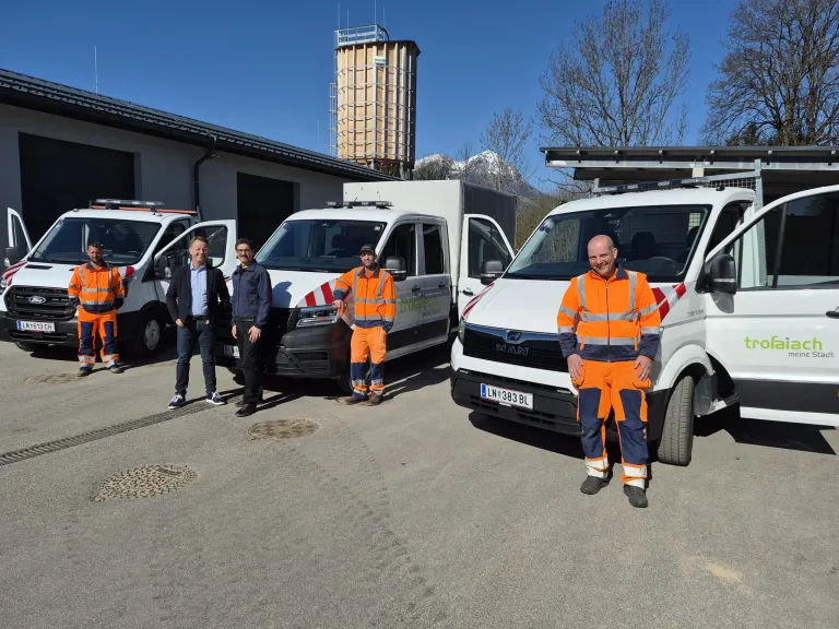 Eine Gruppe von Menschen in orangefarbenen Uniformen steht vor weißen Lieferwagen. Sie alle lächeln. Dahinter befinden sich Gebäude mit einem Silo.