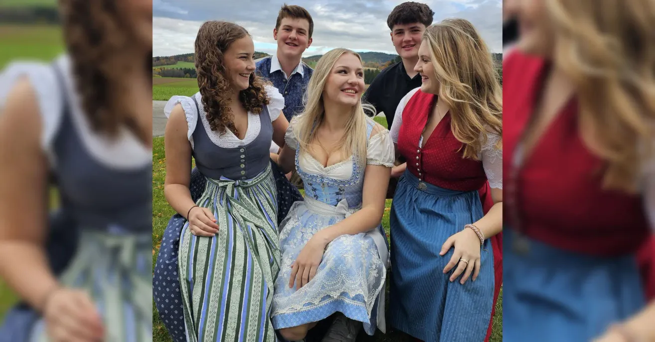 A group of young people dressed in traditional Bavarian clothing are smiling and posing for a photo. The setting is an open field with a cloudy sky.