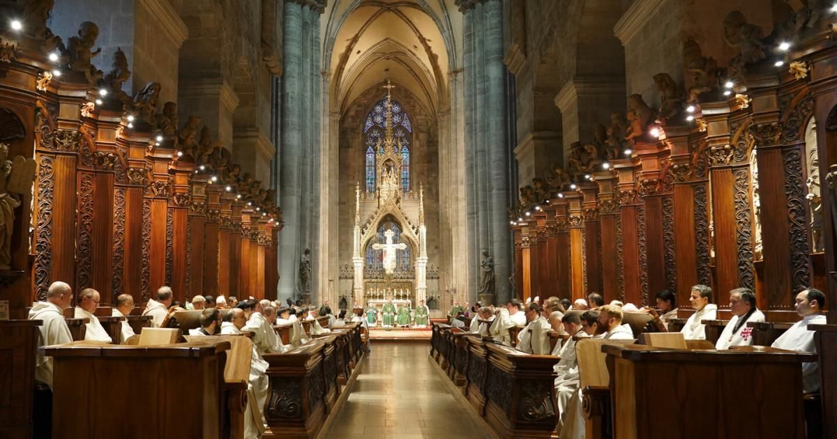 Eine Gruppe von Geistlichen in weißen Gewändern sitzt in den Kirchenbänken einer Kathedrale. Hinter dem Altar befindet sich ein Buntglasfenster.