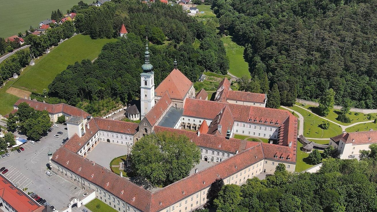 Luftaufnahme eines historischen Gebäudes mit Glockenturm, mehreren Gebäuden und einem breiten Innenhof, umgeben von üppigem Grün und Bäumen.