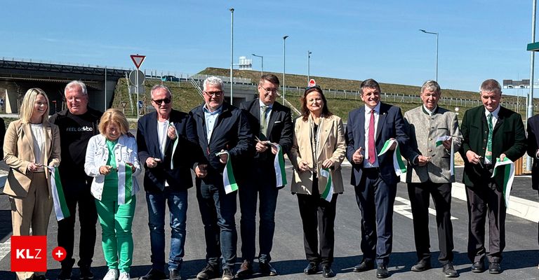 A group of people in suits and ties stand on a road, holding flags and smiling. Behind them is a bridge with street lights.