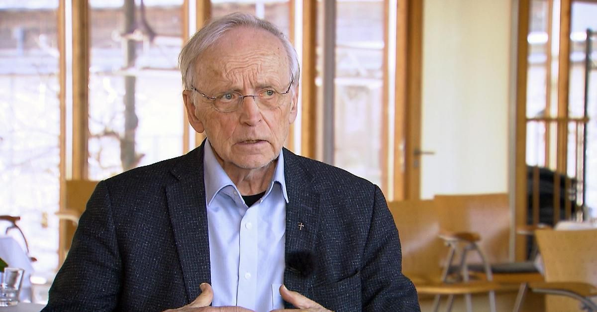 Ein älterer Mann mit Brille und Blazer spricht in einem Raum mit Holzstühlen und Glastüren.