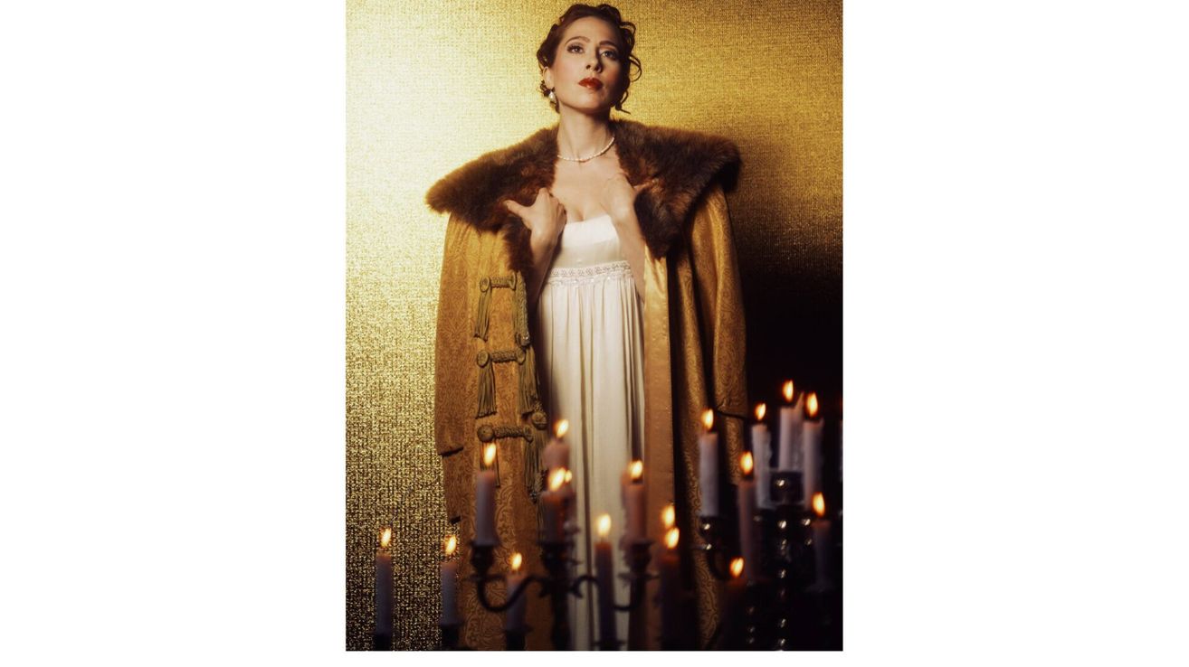 A woman in a white dress and a fur coat is standing in front of a golden wall with candles.