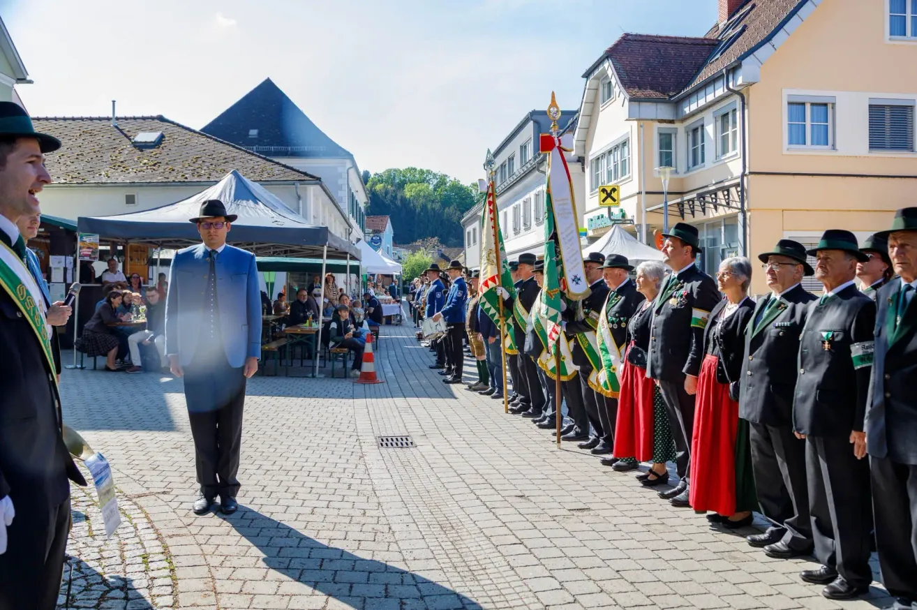 A line of people in traditional attire stand on a brick path, possibly for a parade, with a man in a blue suit standing in front. In the background, there are tents, buildings, and people sitting at tables.