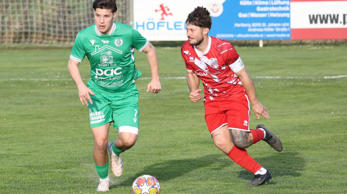 Two soccer players are running on a field. The player in green is looking at the player in red, who is chasing the ball. The ground has grass and some markings. Behind them, a signboard is mounted on the grass.