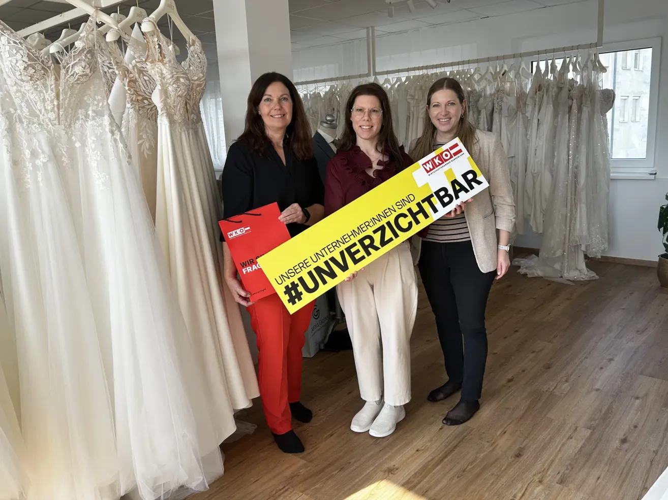 Three women stand in a bridal shop, smiling. One holds a sign that says #Unversichtbar. Behind them, dresses hang on racks.
