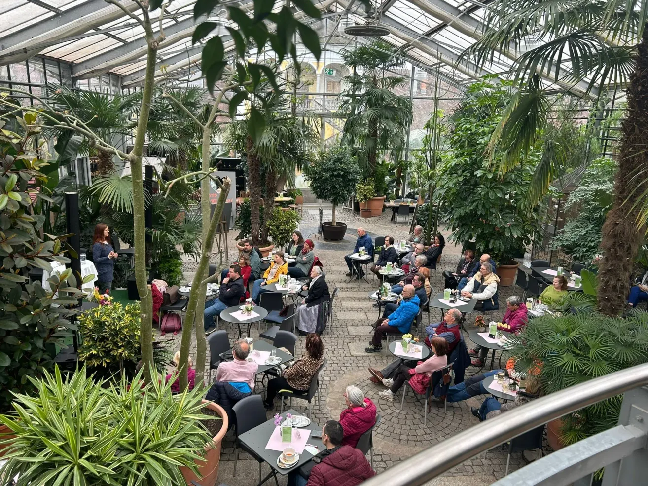 An indoor garden restaurant with glass ceilings, filled with people sitting at tables, surrounded by potted plants. A woman stands on the left, and a man in a blue jacket sits on the right.