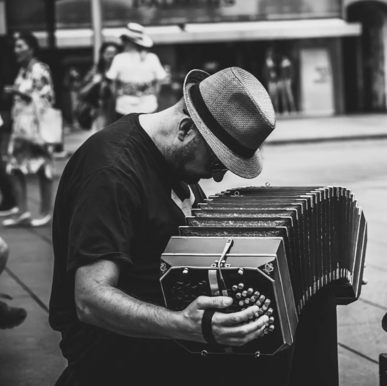 A man in a hat and glasses plays an accordion on a city street. Behind him, people walk by.