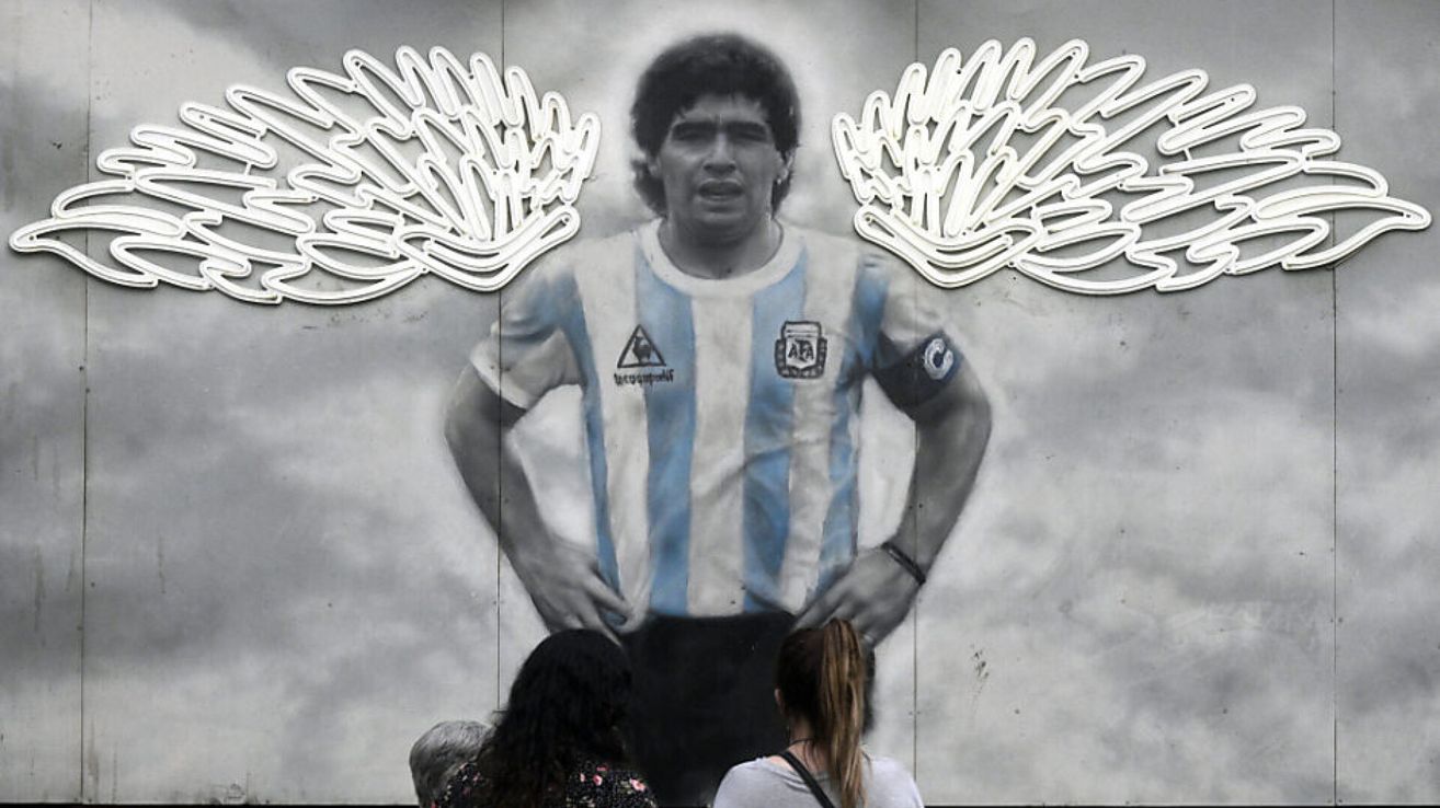 A mural of a soccer player, Diego Maradona, is on a wall. The player wears a white and blue jersey, and two women are looking at him.