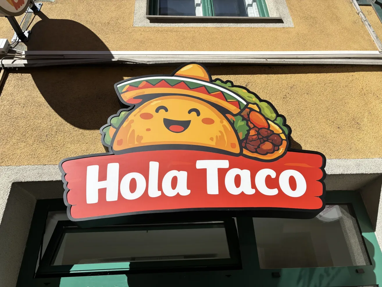 A restaurant sign for Hola Taco shows a smiling taco with a sombrero, placed above a red banner. The restaurant is on a building with a window.