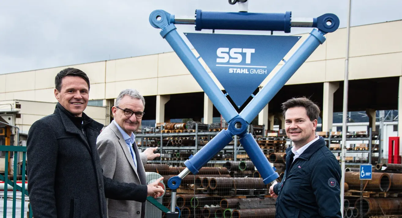 Three men stand in front of a blue metal structure, smiling and looking at the camera. Behind them is a building with a metal structure and pipes. The blue structure has the text 'SST Stahl GmbH'.