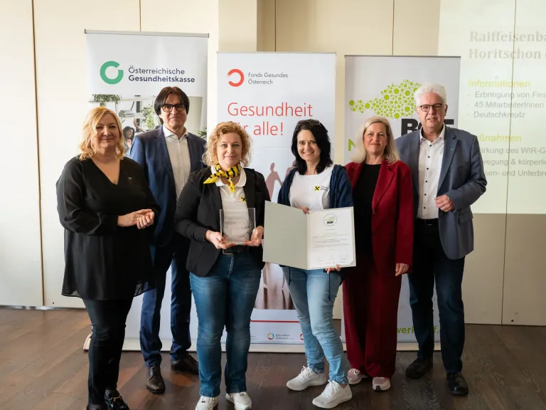 Sechs Personen posieren für ein Bild mit einem Zertifikat und einem Dokument. Eine Frau in der Mitte hält das Dokument. Hinter ihnen sind Banner mit Logos und Text.