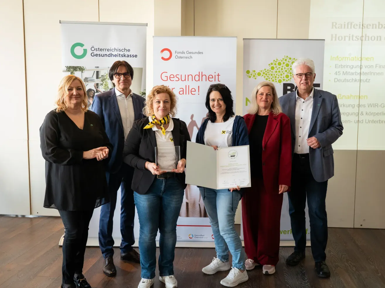 Sechs Personen posieren für ein Bild mit einem Zertifikat und einem Dokument. Eine Frau in der Mitte hält das Dokument. Hinter ihnen sind Banner mit Logos und Text.