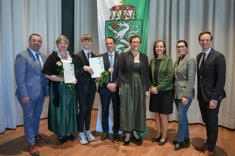 A group of people stand together holding certificates and flowers. They are dressed formally and smiling. A flag with a coat of arms is behind them.