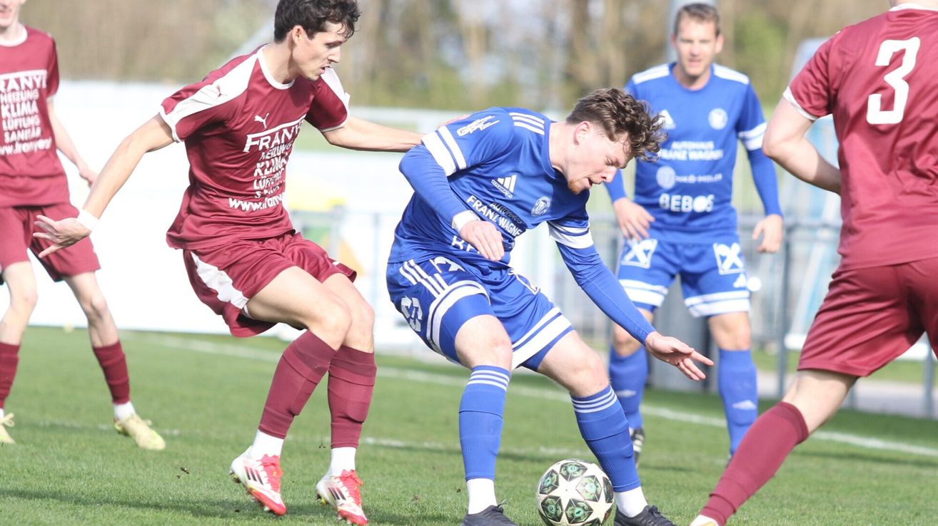 Two soccer players are competing on a field. One in a red uniform and the other in a blue uniform. They are both trying to get the soccer ball.