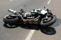 A black and white motorcycle lies on its side on a road, its front wheel detached. Shadows are cast on the road, and there are white markings visible.