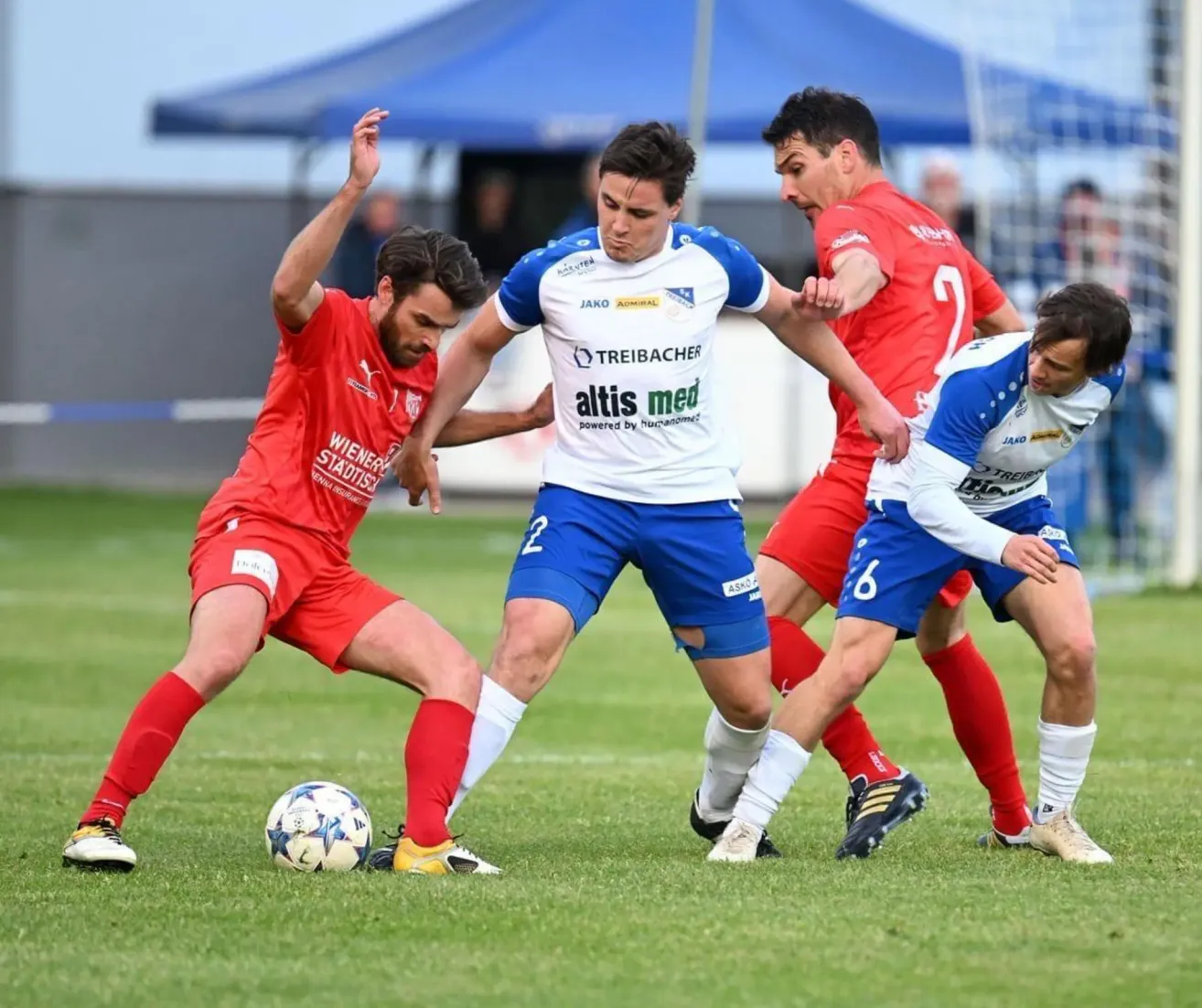 Four soccer players in red and blue jerseys are on a field. They are competing for the ball. The player in red is kicking the ball.