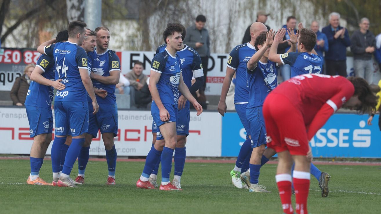 Soccer players in blue uniforms celebrate on the field. Spectators in the background watch the game. A white banner with red letters is behind the players.