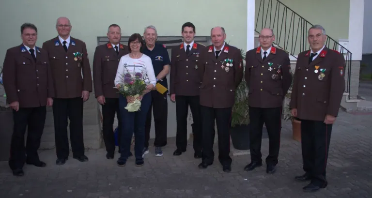 Eine Gruppe von Personen steht draußen; eine Frau hält Blumen, und Männer in Uniformen stehen hinter ihr. Sie alle schauen nach vorne, und ein Gebäude steht hinter ihnen.