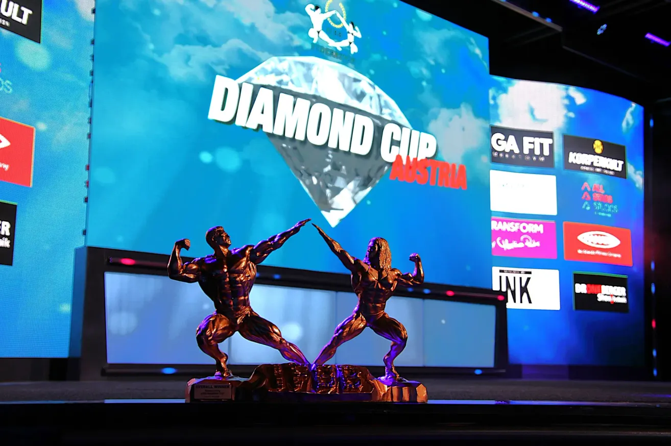 Two bronze statues of muscular men, positioned as if they are flexing their muscles, are displayed on a stage with a large screen showing Diamond Cup Austria in the background.