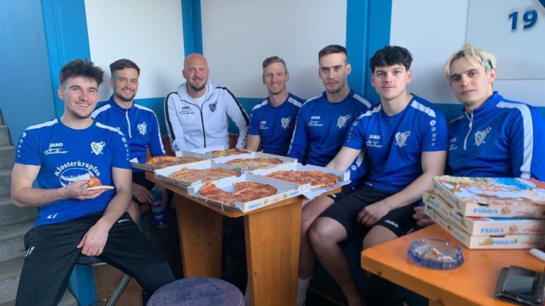 Five men in matching blue jerseys sit around a table with several pizza boxes, smiling and posing for a photo.