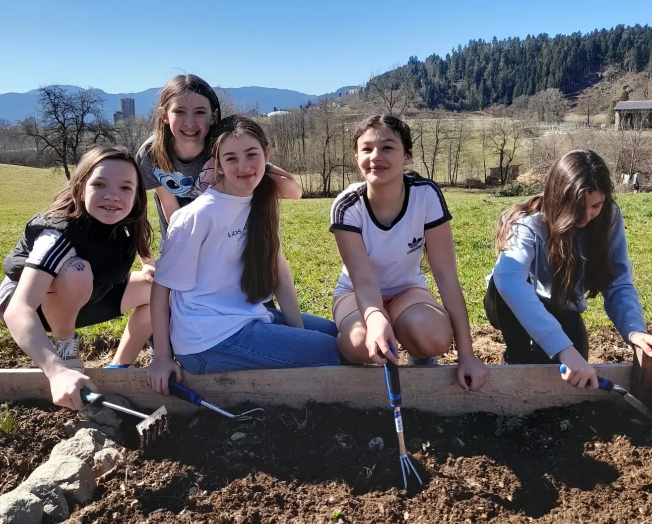 Five girls in a garden, smiling, holding gardening tools, sitting on a wooden bed. Behind them are mountains and trees.